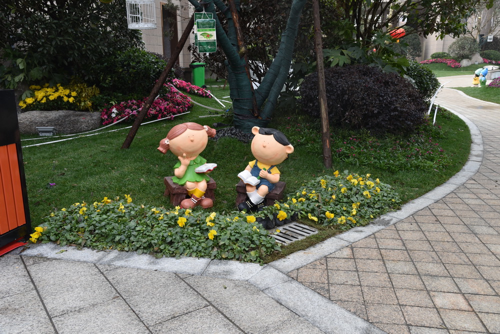 天门碧桂园·天悦府示范区实景展示绿油油的草地，胜开的鲜花，两个卡通人物坐在此学习，增加活泼感